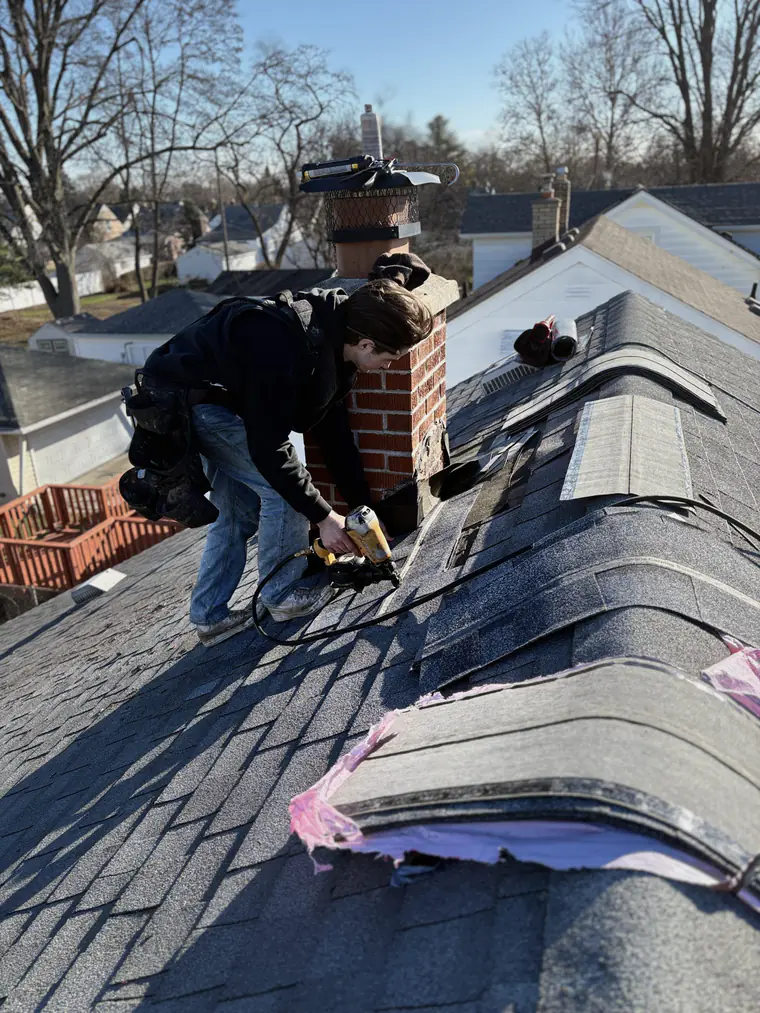 A Joe’s Roofing crew member nailing ridge cap shingles around a brick chimney.